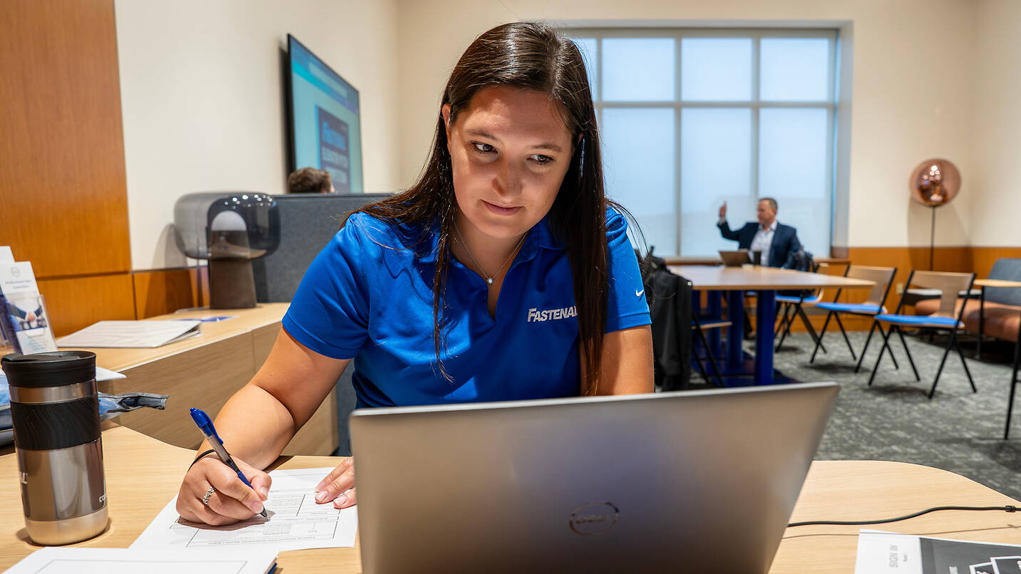 A person in a blue polo shirt with "Fastenal" embroidered on it is seated at a desk, writing on paper while looking at a laptop.